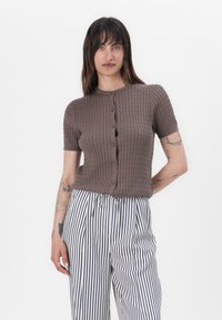 Donna con lunghi capelli scuri indossa una maglia a maniche corte marrone con bottoni e pantaloni bianchi a vita alta con strisce verticali nere, in piedi davanti a uno sfondo bianco.