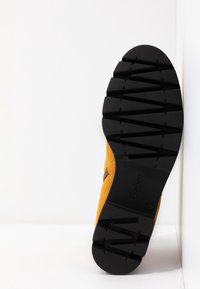 Botte en daim avec un dessus jaune moutarde et une semelle en caoutchouc noir texturé. Présente un motif de semelle robuste pour une bonne adhérence et durabilité.