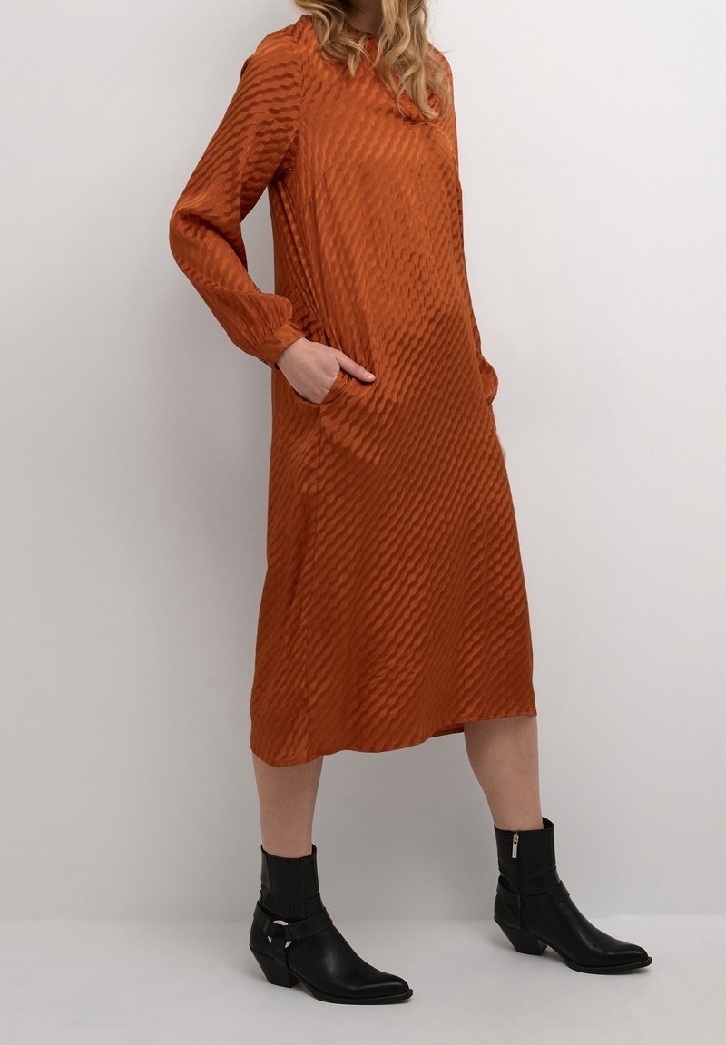 Femme portant une robe midi orange texturée à manches longues avec poches latérales et bottines noires, debout contre un mur uni.