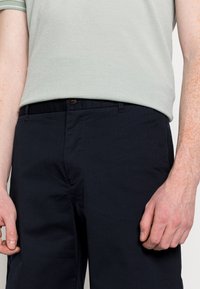 Pantalons en coton bleu marine avec une texture lisse, dotés d'une fermeture à bouton et d'un design à plat. Portés avec un polo vert clair.
