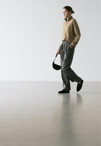 Pull beige en maille torsadée avec col zippé, pantalon plissé gris et chaussures noires sans lacets, tenant un petit sac noir. Fond blanc.