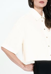 Cropped, short-sleeve blouse in light cream fabric with a button front. Features a classic collar and subtle sheen, paired with black pants.