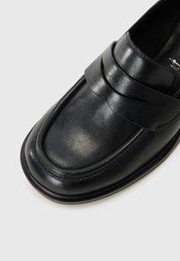 Svart läderloafersko med rund tå, som har ett horisontellt band över ovansidan och subtila sömnadsdetaljer. Slät yta.
