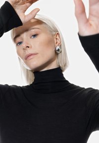 Maglione nero a collo alto con texture a righe. Orecchini rotondi in argento. Capelli chiari, tratti marcati, occhi blu, mano sollevata contro uno sfondo bianco.