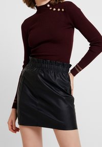 Haut à manches longues côtelé bourgogne avec des accents de boutons dorés, associé à une jupe en cuir synthétique noire avec une taille élastique.