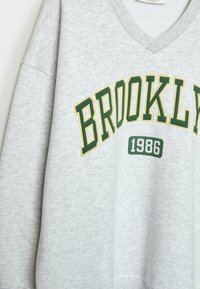 Grå sweatshirt med V-ringning, med "BROOKLYN" i grön och gul brodering och året "1986" i grönt nedanför.