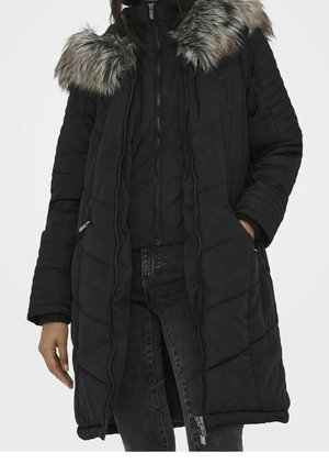 Femme portant un long manteau d'hiver matelassé noir avec une capuche doublée de fourrure, fermeture éclair partiellement ouverte, mains dans les poches, associé à un jean noir.
