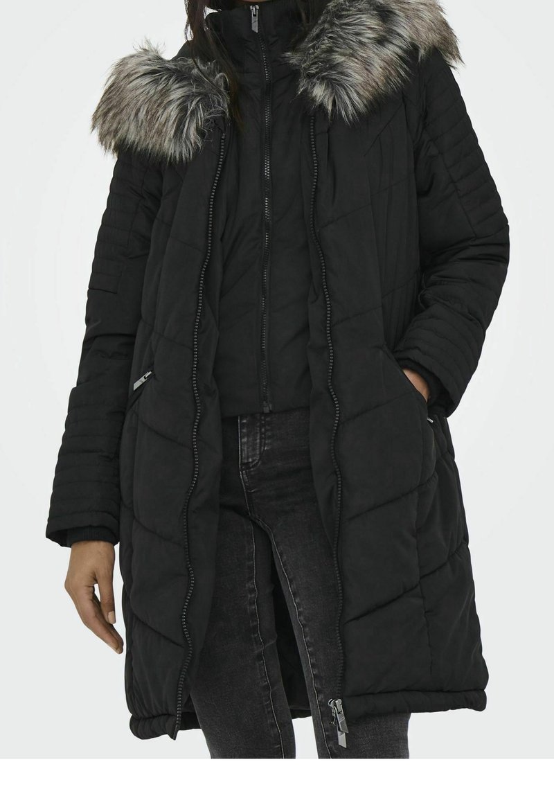 Femme portant un long manteau d'hiver matelassé noir avec une capuche doublée de fourrure, fermeture éclair partiellement ouverte, mains dans les poches, associé à un jean noir.