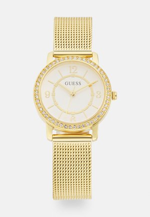 Gold mesh strap watch with a round case. White dial features gold hour markers and crystal accents. Brand name "GUESS" displayed.