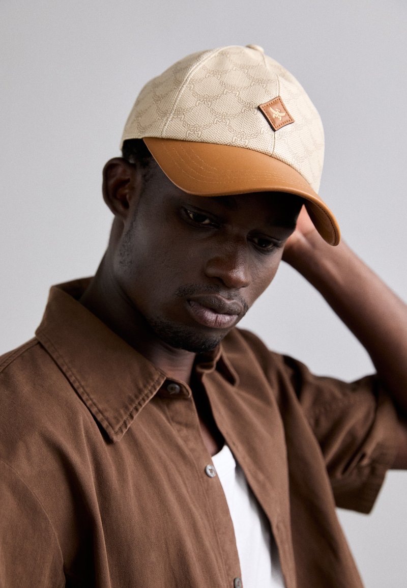 Casquette beige avec un dessus en tissu à motifs et un bord brun. Présente un patch en simili cuir à l'avant. Le mannequin porte une chemise marron à boutons.