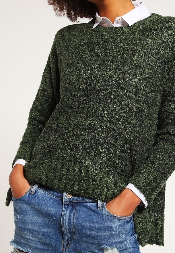 Personne portant un pull en maille texturé vert foncé sur une chemise blanche à col, les mains dans les poches d'un short en jean bleu déchiré.