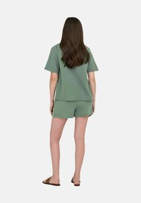 Top corto a maniche corte di tessuto verde con pantaloni abbinate. Vestibilità comoda, dettagli di cuciture a vista e orli piatti. Indossato con sandali marroni.
