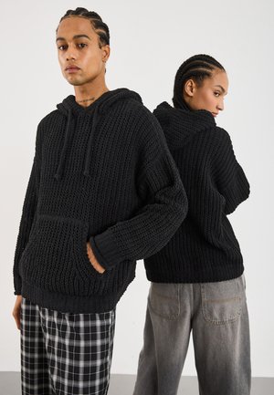 Zwarte gebreide hoodie met een textuurpatroon, voorzien van een voorzak, trekkoorden en een losse pasvorm, gecombineerd met geruite en grijze broek.