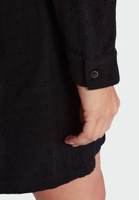 Acercamiento a una manga de vestido de tela negra texturizada con un botón negro bordeado de oro en el puño, y una mano reposando junto al dobladillo del vestido.