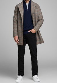 Cappotto a quadri marrone con una vestibilità su misura, indossato sopra un maglione blu navy con zip, pantaloni neri e sneakers bianche. Tessuto morbido e linee pulite.