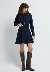 Granatowa sukienka z guzikami, z zamkiem z przodu, paskiem w talii i rozkloszowanym dołem. Połączona z brązowymi skórzanymi kozakami do kolan. Gładka tekstura.