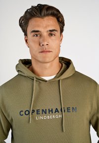 Olivgrön hoodie tillverkad av mjukt tyg, med en frontrik ficka, justerbar dragsko och tryckt "COPENHAGEN"-logotyp i mörkblått.