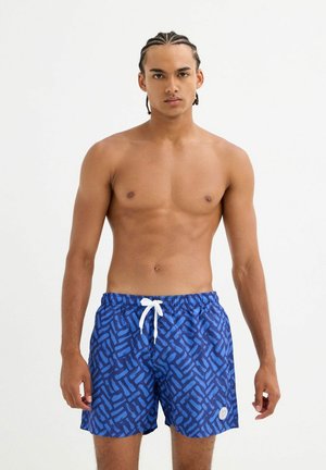 Joven con el cabello trenzado, vistiendo pantalones cortos de baño azules con estampado y cordón blanco, de pie frente a un fondo blanco liso.