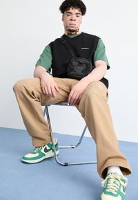 Čierna a zelená košeľa s krátkym rukávom, béžové nohavice, čierna crossbody taška a zelené tenisky Nike s bielymi akcentmi. Model sedení na stoličke.