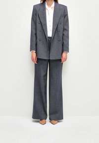 Blazer gris de espiga de doble botonadura combinado con pantalones de pierna ancha a juego. Camisa blanca por debajo y zapatos de punta marrón.