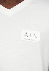 Biele tričko s V-krkom vyrobené z hladkej tkaniny, ktoré má zvýšenú striebornú nášivku s textom "A|X Armani Exchange" na ľavej strane hrude.