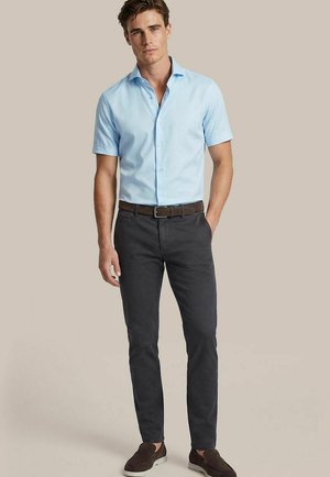 Mann trägt hellblaues, kurzärmeliges Hemd mit Knöpfen, dunkle, eng geschnittene Hose, braunen Gürtel und braune Slipper, steht vor beigem Hintergrund.