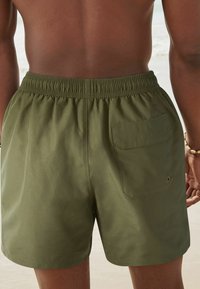 Olívazöld fürdőnadrág, rugalmas derékpánttal, hátsó zsebbel és sima, könnyű anyagból készült. Nincsenek látható minták vagy díszítések.