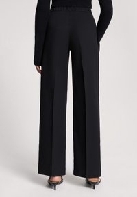 Osoba w czarnych, szerokich spodniach z wysokim stanem, czarnej bluzce z długim rękawem i czarnych sandałach na obcasie, stojąca na jednolitym tle.