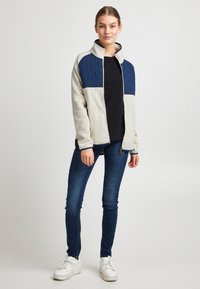 Krēmkrāsas vilnas jaka ar tumši ziliem šūtiem akcentiem, rāvējslēdzēja aizdare, augsta apkakle, ribota aproce; tiek valkāta kopā ar tumši ziliem džinsiem un baltām sporta kurpēm.