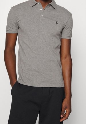 Man wearing a fitted grey polo shirt with two buttons and black embroidered horse logo on chest, paired with black pants.