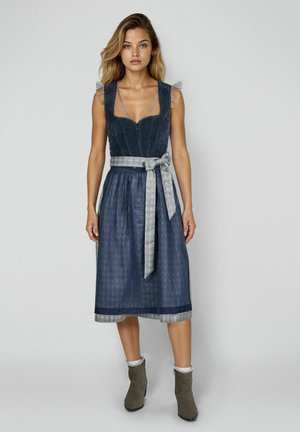 Frau trägt ärmelloses dunkelblaues Dirndl mit Spitzendetails und gemustertem Gürtel, kombiniert mit grauen Knöchelstiefeln und Spitzenstrümpfen.