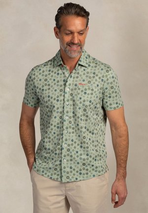 Man draagt een lichtgroen overhemd met korte mouwen, met blauwe en bruine bladmotieven, en beige broek, handen in de zakken, met een lichte glimlach.