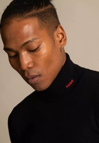 Black turtleneck sweater with a red "HUGO" logo on the collar, featuring a fitted design and smooth texture. Model has short hair and an earring.