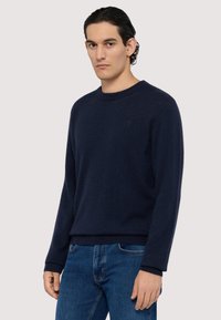 Mörkblå stickad tröja med rund halsringning, långa ärmar och ribbade muddar. Strukturerad tyg med en diskret logodetalj på bröstet.