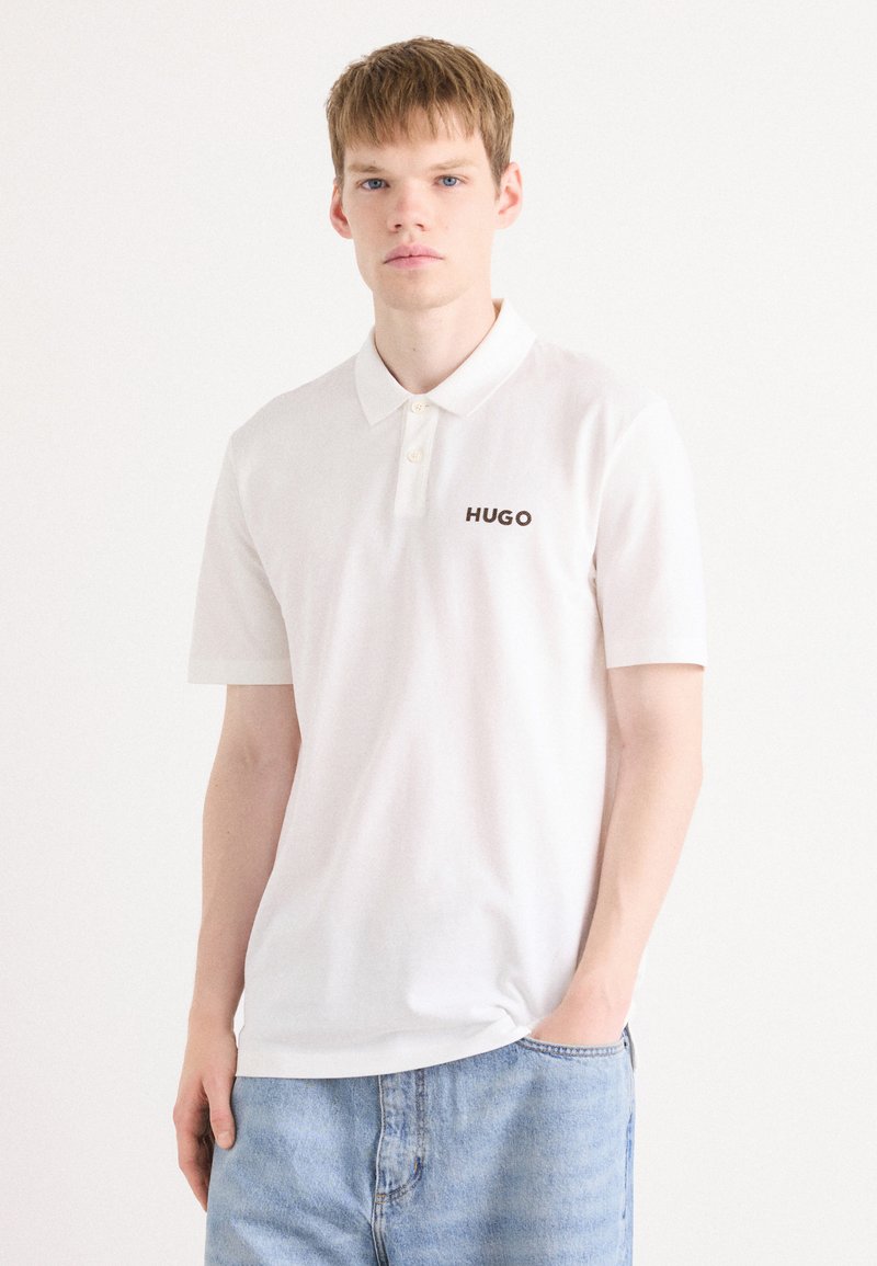 Hvit polo skjorte med krage og tre knapper, med en kontrastfarge svart "HUGO" logo på venstre bryst. Laget av bomull, kortermet.