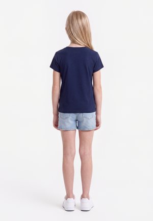 Jong meisje met lang blond haar, staand met de rug naar de kijker, draagt een marineblauw T-shirt, lichte denimshorts en witte sneakers.