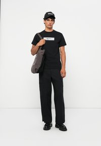 Černé tričko s logem, černé cargo kalhoty a tmavě šedá taška. Celý outfit doplňuje černá kšiltovka. Jednoduchý, funkční design.