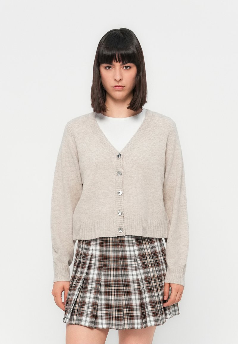 Cardigan beige lavorato a maglia con scollo a V, chiusura con bottoni e maniche lunghe, abbinato a una gonna plissettata a quadri in tonalità di marrone e bianco.
