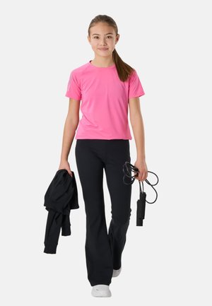 Ung pige iført lyserød skjorte og sorte bukser, der holder en sort jakke i den ene hånd og et sjippetov i den anden, går fremad på hvid baggrund.