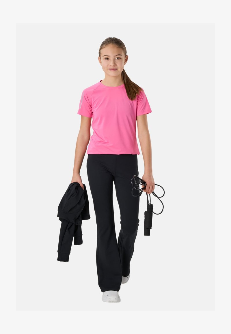 Jeune fille portant un t-shirt rose et un pantalon noir, tenant une veste noire dans une main et une corde à sauter dans l'autre, marchant vers l'avant sur fond blanc.