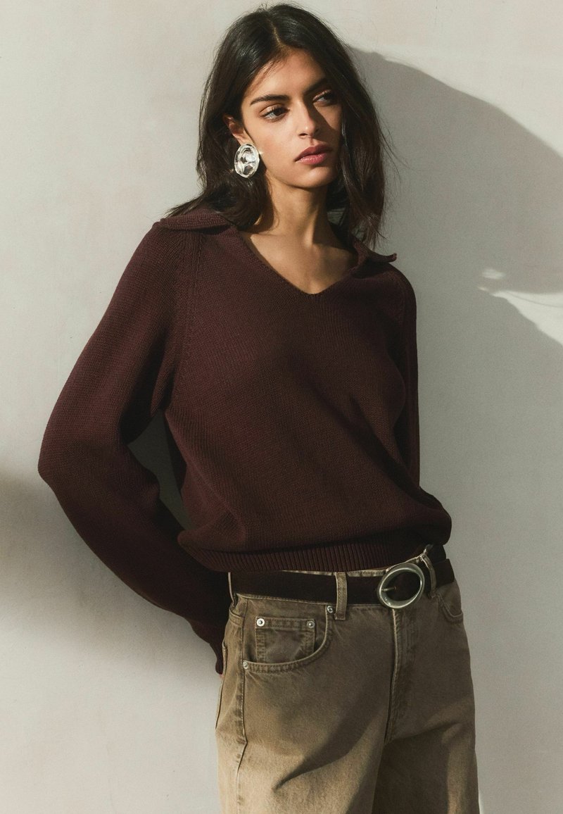 Žena oblečená v tmavohnedom svetri s výstrihom do V, béžových džínsoch s veľkou okrúhlou opaskovou prackou a priehľadnými veľkými kruhovými náušnicami, stojaca opretá o stenu.