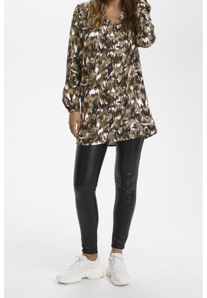 Frau trägt eine langärmelige Tunika mit abstraktem Muster, schwarze Leggings aus Kunstleder und weiße klobige Sneaker vor weißem Hintergrund.