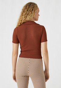 Ein rostfarbener Kurzarm-Oberteil mit gerippter Textur und Kragen, kombiniert mit einer engen Hose mit braun-creamfarbigem Karomuster.