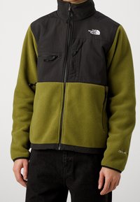 Groene en zwarte fleecejas met een hoge kraag, twee borstzakken en ritssluitingdetails. Zachte textuur en contrasterende kleurvlakken.