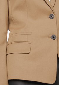 Blazer marrone con un design elegante, caratterizzato da due tasche frontali, dettagli con bottoni e un tessuto a trama. Vestibilità su misura e linee pulite.