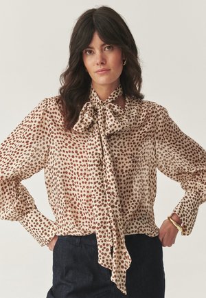 Vrouw met donker golvend haar, gekleed in een beige blouse met bruine stippen en een grote strik, gecombineerd met donkere broek en een gouden armband.