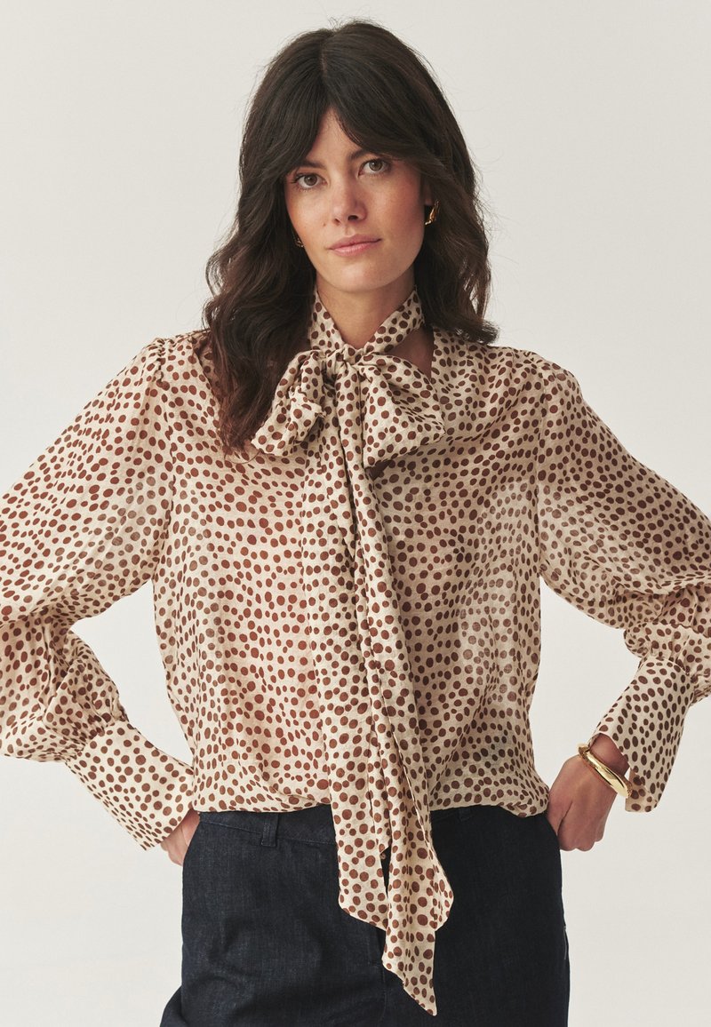 Femme aux cheveux ondulés foncés portant un chemisier beige à pois marron et un grand nœud, associé à un pantalon sombre et un bracelet en or.