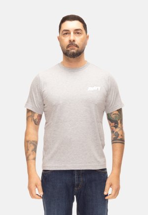Hombre con cabello corto y barba, vestido con una camiseta gris clara y vaqueros azules, de pie con tatuajes visibles en ambos antebrazos.