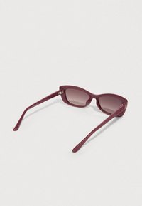 Γυαλιά ηλίου σε μπορντό χρώμα με σχήμα cat-eye, κατασκευασμένα από γυαλιστερό πλαστικό. Διαθέτουν φακούς με διαβάθμιση χρώματος και υφασμένους βραχίονες.