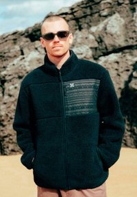 Hombre con una chaqueta de forro polar de estampado oscuro y gafas de sol está de pie con las manos en los bolsillos frente a un fondo rocoso al aire libre.